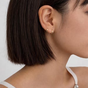 Puede incluir: Primer plano de una oreja con dos pequeños pendientes de aro dorados. Los pendientes tienen un diseño sencillo y clásico, que refleja la luz. La persona tiene el pelo corto y castaño oscuro y lleva una camiseta blanca de tirantes.