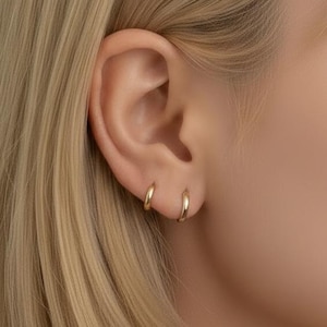 Op de afbeelding: Close-up van een oor met twee kleine, gouden oorringen. De oorbellen hebben een eenvoudig, klassiek ontwerp met een gladde, gepolijste afwerking. Het oor is geplaatst tegen een achtergrond van blond haar.