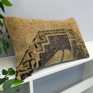 Puede incluir: Un cojín rectangular de tela tejida con textura en tonos bronceado y beige. El cojín muestra un patrón geométrico prominente en gris carbón y verde oliva, caracterizado por líneas angulares y formas simétricas en su diseño.