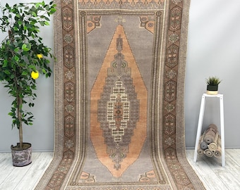 Puede incluir: Una alfombra rectangular grande con un intrincado diseño geométrico. La alfombra presenta una forma de diamante central en tonos melocotón y gris, rodeada por un borde con patrones florales y geométricos. La alfombra se exhibe sobre un suelo de madera clara.