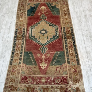 May include: A rectangular, distressed rug with a central red field, framed by a beige border. The rug features geometric patterns in shades of green, beige, and brown. The rug's design includes a central medallion and corner accents.