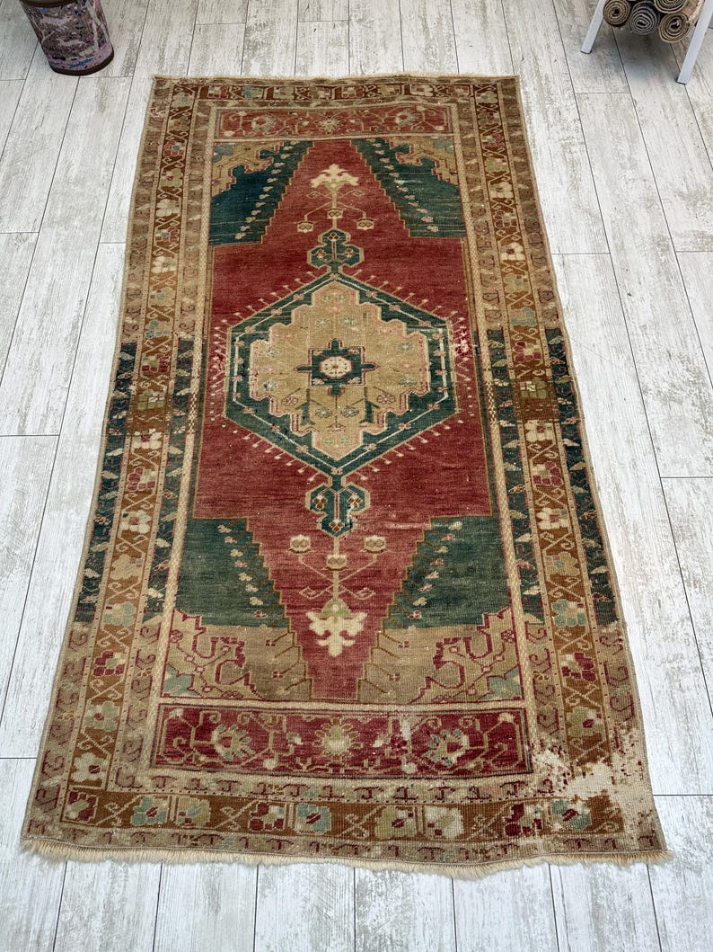 May include: A rectangular, distressed rug with a central red field, framed by a beige border. The rug features geometric patterns in shades of green, beige, and brown. The rug's design includes a central medallion and corner accents.