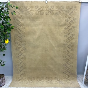 May include: A large, rectangular rug in a muted tan color. The rug features a detailed border design in a darker shade of tan, with repeating floral and geometric patterns. The rug is hanging, showing its full size and texture.
