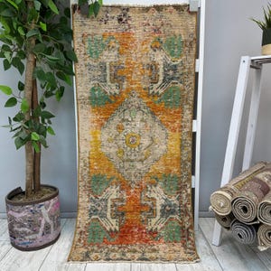 May include: A rectangular runner rug featuring a distressed, weathered texture with a central diamond medallion. The color palette includes vibrant shades of orange, mustard yellow, teal, and charcoal gray, finished with short fringe at both ends.