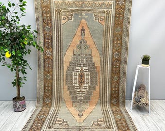 Puede incluir: Una alfombra rectangular grande con un diseño central de diamante en tonos melocotón, gris y marrón. La alfombra presenta intrincados patrones geométricos y un borde con motivos florales repetidos. La alfombra se exhibe sobre un fondo gris neutro.
