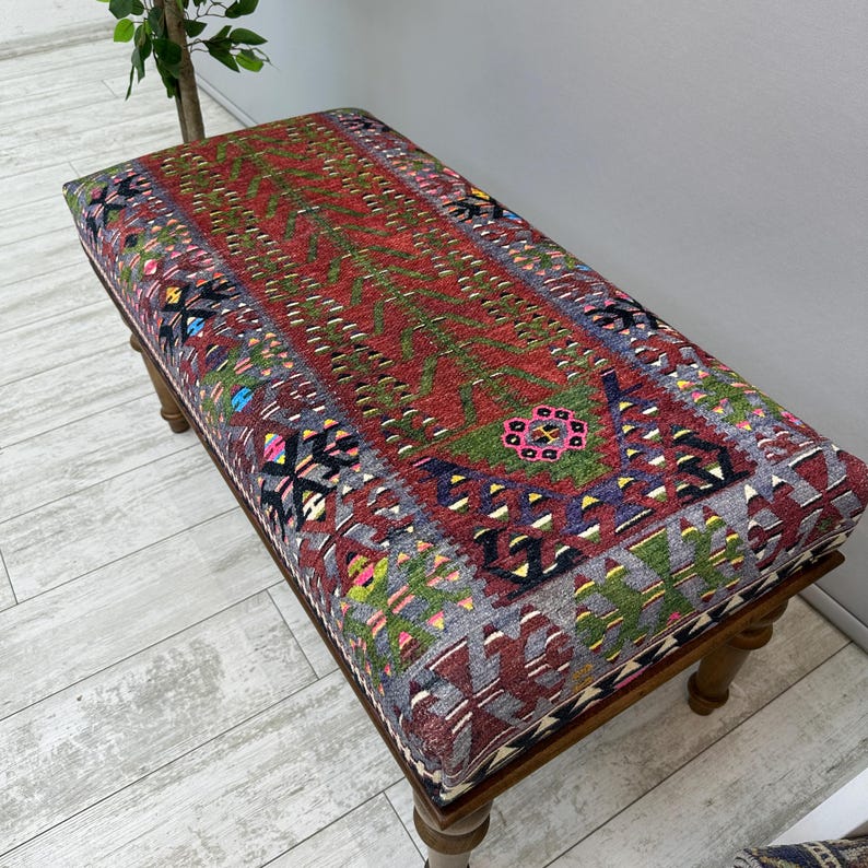 May include: A rectangular upholstered bench with a detailed, colorful rug-like pattern. The bench has a wooden frame and legs. The rug design features red, green, gray, and other colors. The bench is a decorative furniture piece.