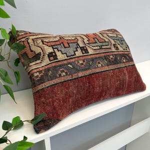 Op de afbeelding: Rechthoekig decoratief kussen met een vintage patroon. Het bovenste gedeelte heeft een geometrisch patroon in beige, oranje, grijs en groen. Het onderste gedeelte is effen dieprood. Het kussen ligt op een wit oppervlak.