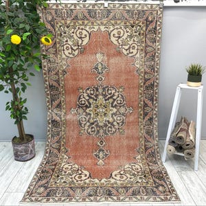 Puede incluir: Una alfombra vintage con un fondo melocotón descolorido y intrincados patrones florales en tonos marrones, crema y negro. La alfombra tiene un aspecto desgastado y un borde deshilachado.