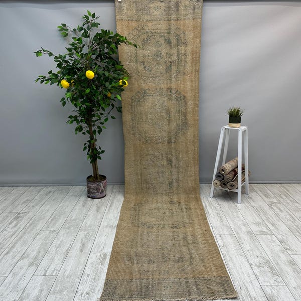 May include: A long, beige runner rug with a faded, distressed pattern. The rug has a subtle green and brown colour scheme and a fringed edge.