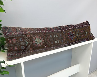 Puede incluir: Un cojín lumbar largo y rectangular que presenta un tejido oscuro y multicolor con intrincados patrones geométricos en tonos marrón, rojo oscuro, azul y crema. La tela tiene una apariencia texturizada y envejecida con un diseño de motivos repetitivos.
