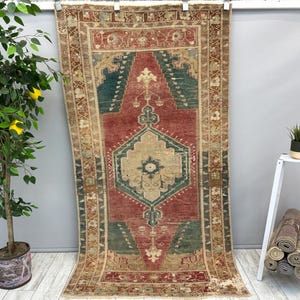 May include: A rectangular, vintage-style rug with a central design in red, teal, and beige. The rug features an intricate geometric pattern with a border in shades of brown and beige. The rug is displayed hanging against a gray wall.