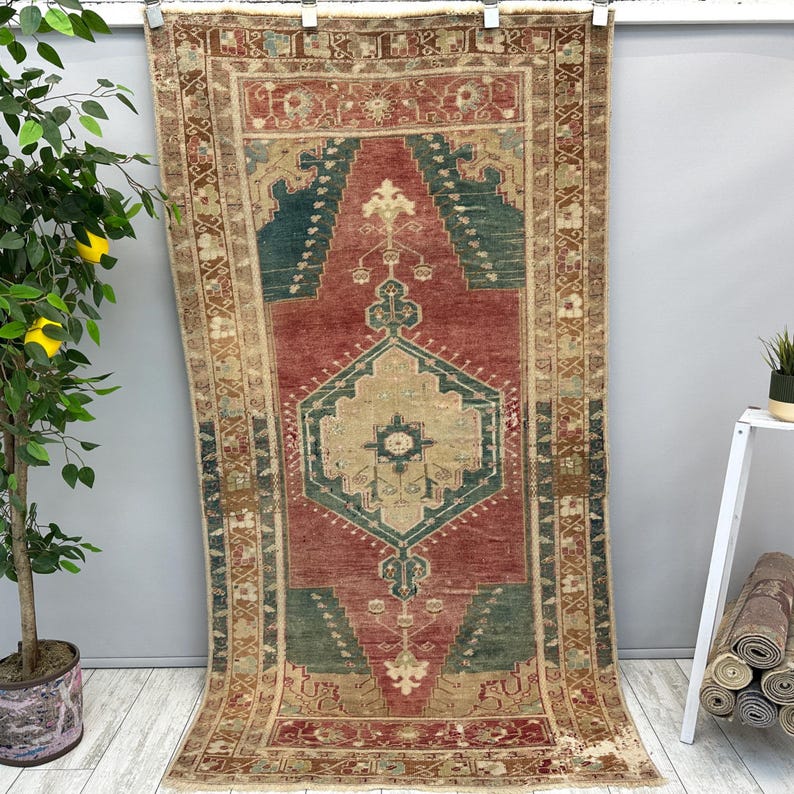 May include: A rectangular, vintage-style rug with a central design in red, teal, and beige. The rug features an intricate geometric pattern with a border in shades of brown and beige. The rug is displayed hanging against a gray wall.