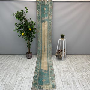 May include: A long, narrow runner rug featuring a central beige panel flanked by wide teal borders. The borders are decorated with repeating geometric patterns and floral motifs in muted tones. The rug has a weathered, vintage appearance.