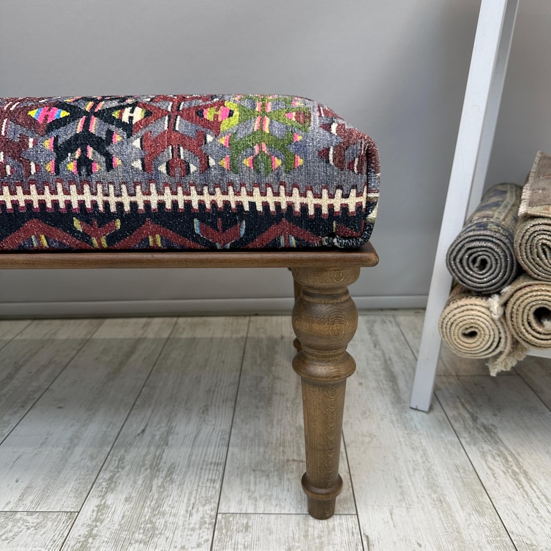 May include: A rectangular ottoman with a patterned top and wooden legs. The top features a geometric design in shades of red, gray, black, and yellow. The wooden legs are a warm brown color.