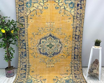 Puede incluir: Una alfombra rectangular grande con una apariencia vintage y desgastada. La alfombra presenta un campo central amarillo dorado con un medallón grande y ornamentado en azul marino y turquesa. El borde es azul marino con patrones florales. La alfombra se exhibe contra una pared neutra.