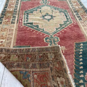 May include: A distressed, rectangular rug with a central geometric design in beige, teal, and red. The rug's border features intricate patterns in shades of brown and beige, with a portion of the rug folded over, revealing the underside.