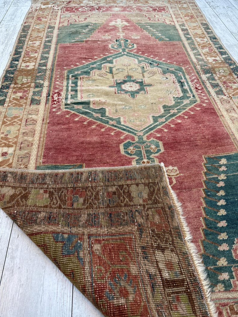 May include: A distressed, rectangular rug with a central geometric design in beige, teal, and red. The rug's border features intricate patterns in shades of brown and beige, with a portion of the rug folded over, revealing the underside.