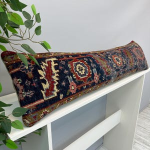 May include: A long, rectangular lumbar pillow featuring a dark navy blue base with intricate, multi-colored geometric and floral patterns in shades of red, cream, and muted green. The textile has a textured, woven appearance with visible pile.