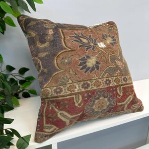 Puede incluir: Un cojín cuadrado con una funda de tejido con un complejo patrón floral y geométrico en tonos rojo intenso, marrón oscuro, azul apagado y beige. El diseño incluye motivos botánicos intrincados y un borde decorativo.