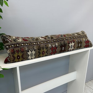 May include: A long, rectangular lumbar pillow featuring a woven textile cover with a muted taupe background. The surface displays a repeating geometric pattern in shades of deep red, olive green, cream, and dark brown, with a central horizontal border.