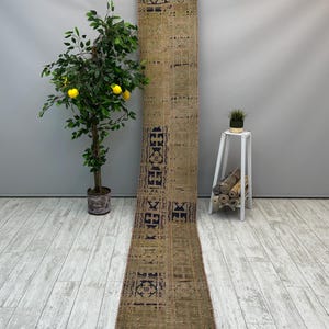 May include: A long, narrow, green and brown rug with a geometric pattern in black. The rug is hanging on a wall with a small potted plant and a white stand with a potted plant on the right side of the image.