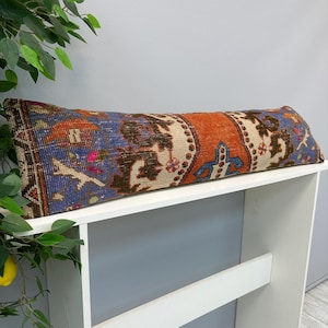 May include: A long rectangular lumbar pillow featuring a woven textile pattern with vibrant shades of orange, blue, brown, and cream. The fabric displays intricate geometric and floral motifs characteristic of traditional rug-style weaving techniques.