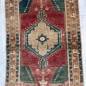 May include: A rectangular, vintage rug with a central geometric design in shades of red, green, and beige. The rug features intricate patterns and borders, showcasing a traditional aesthetic. The rug's dimensions are not visible.