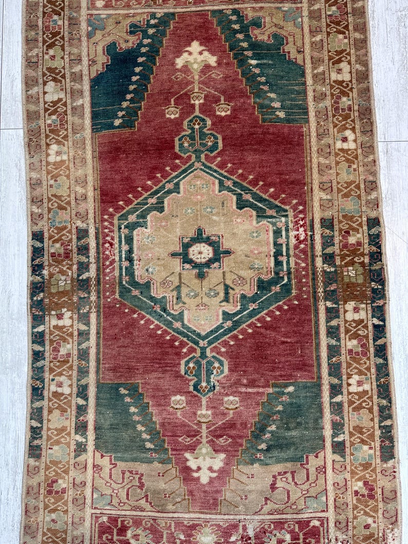 May include: A rectangular, vintage rug with a central geometric design in shades of red, green, and beige. The rug features intricate patterns and borders, showcasing a traditional aesthetic. The rug's dimensions are not visible.