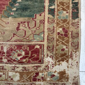 May include: Close-up of a section of a vintage rug with a distressed appearance. The rug features a complex pattern in shades of burgundy, teal, beige, and brown. The edges are frayed, and the rug has a worn, antique aesthetic.