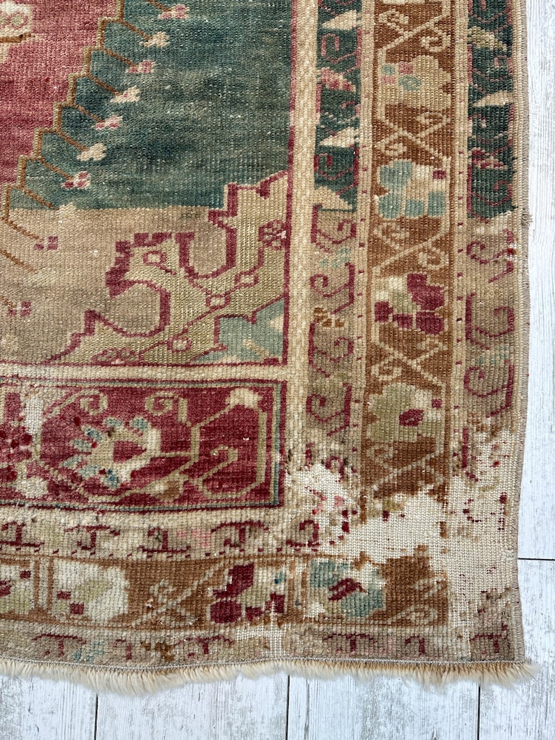 May include: Close-up of a section of a vintage rug with a distressed appearance. The rug features a complex pattern in shades of burgundy, teal, beige, and brown. The edges are frayed, and the rug has a worn, antique aesthetic.
