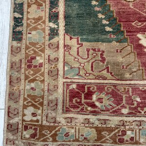 May include: Close-up of a section of a vintage rug with a distressed appearance. The rug features a color palette of red, green, beige, and brown. The design includes geometric and floral patterns, with a fringed edge. The rug is placed on a white wooden surface.