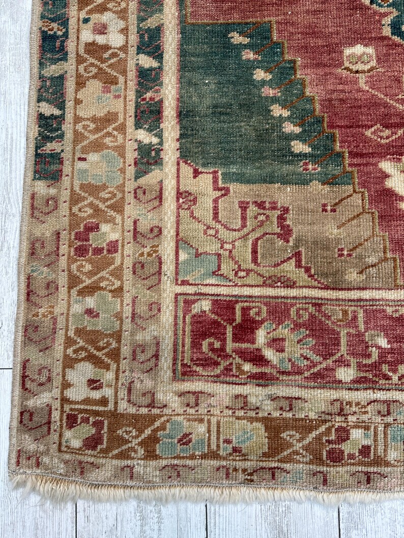 May include: Close-up of a section of a vintage rug with a distressed appearance. The rug features a color palette of red, green, beige, and brown. The design includes geometric and floral patterns, with a fringed edge. The rug is placed on a white wooden surface.
