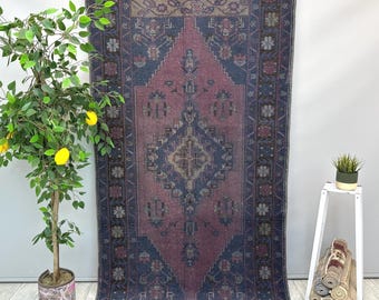 May include: A rectangular, vintage-style rug with a central diamond pattern in shades of mauve and navy blue. The rug features intricate floral and geometric designs along the borders and within the central field. The rug is displayed against a neutral wall.
