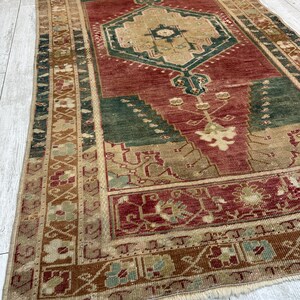 May include: A rectangular, vintage rug with a central geometric design in shades of red, green, and beige. The rug features intricate patterns and a fringed edge. The rug's dimensions are approximately 60 inches by 90 inches.
