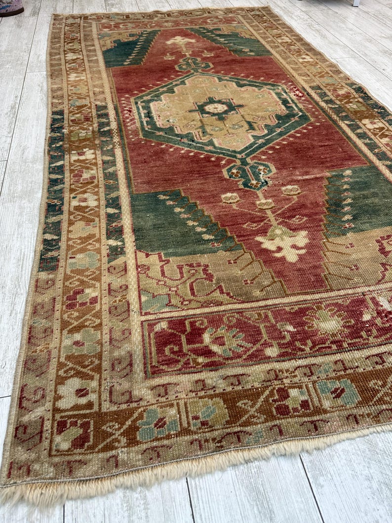 May include: A rectangular, vintage rug with a central geometric design in shades of red, green, and beige. The rug features intricate patterns and a fringed edge. The rug's dimensions are approximately 60 inches by 90 inches.