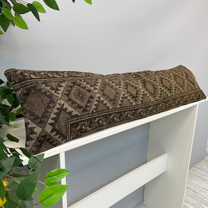 Puede incluir: Un cojín largo y rectangular con un patrón geométrico marrón y beige. El cojín se muestra en una estantería blanca, resaltando su diseño detallado. El patrón presenta formas de diamantes y zigzags.