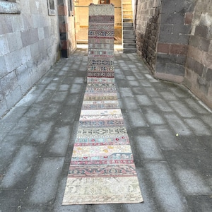 May include: A long, narrow, vintage rug with a patchwork design. The rug features a variety of colors including red, blue, beige, and green, with geometric and floral patterns. The rug is displayed outdoors on a stone surface.
