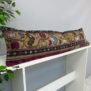 May include: A long, rectangular lumbar pillow featuring a woven textile pattern with a tan base and intricate floral motifs in shades of magenta, blue, yellow, and brown. The pillow has a dark border with a geometric pattern and a deep magenta edge.