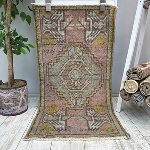 Può includere: Un tappeto rettangolare vintage con un motivo geometrico rosa e grigio. Il tappeto presenta una forma a diamante centrale e motivi ripetuti in giallo e marrone. Il tappeto è esposto su uno sfondo bianco.