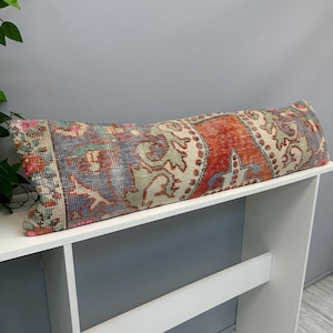 May include: A long rectangular lumbar pillow featuring a woven textile pattern with a distressed appearance. The design incorporates a vibrant color palette of burnt orange, muted blue, sage green, and cream, displaying intricate floral motifs.