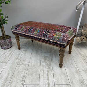 May include: A rectangular bench with a patterned top in shades of red, purple, and green. The bench has turned wooden legs and a dark border. The patterned top has a geometric design. The bench is a decorative furniture piece.
