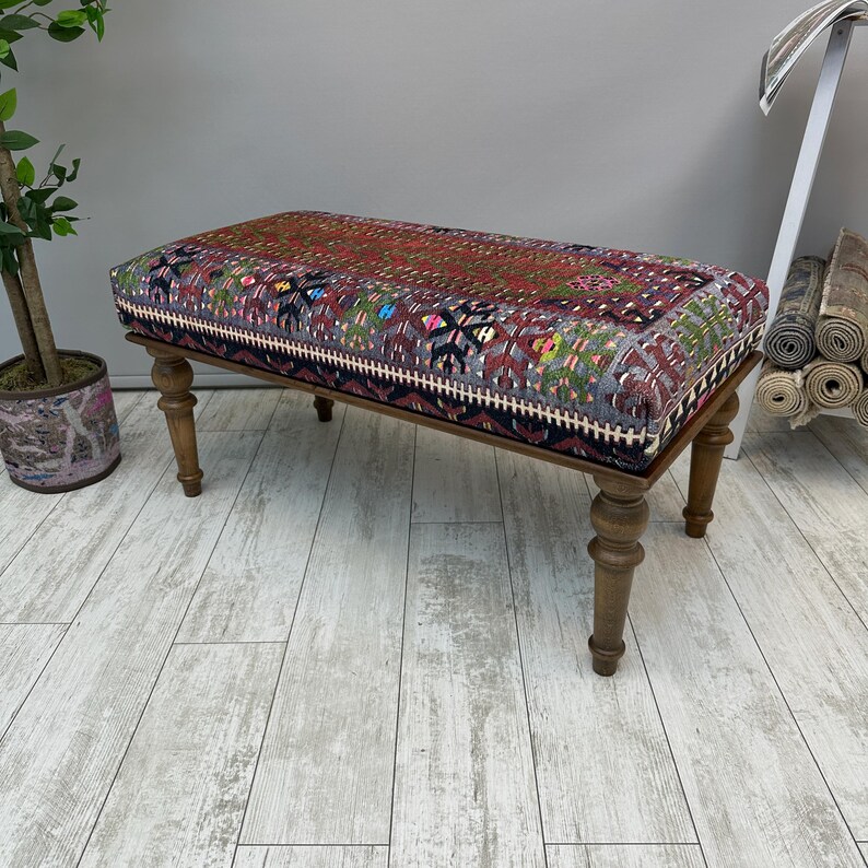 May include: A rectangular bench with a patterned top in shades of red, purple, and green. The bench has turned wooden legs and a dark border. The patterned top has a geometric design. The bench is a decorative furniture piece.