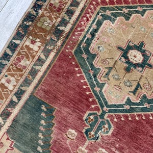 May include: Close-up of a vintage rug with a geometric design. The rug features a central field of deep red, bordered by teal and tan patterns. The intricate details include floral and geometric motifs.