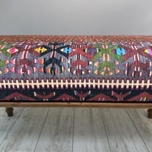 May include: A low wooden bench with a colorful, patterned cushion. The cushion features a geometric design in shades of red, blue, green, and yellow on a gray background. The bench has four short, rounded legs.