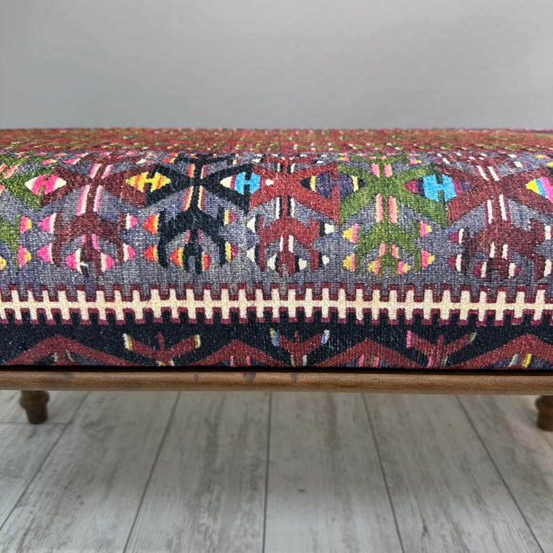 May include: A low wooden bench with a colorful, patterned cushion. The cushion features a geometric design in shades of red, blue, green, and yellow on a gray background. The bench has four short, rounded legs.