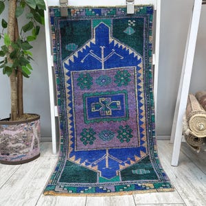 Puede incluir: Una alfombra tejida rectangular que presenta un campo central morado con motivos geométricos verdes, rodeado por un borde azul vibrante y secciones exteriores verde oscuro. El diseño incluye patrones complejos en tonos azul, beige y cerceta.