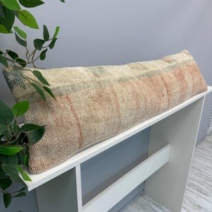 May include: A long, rectangular lumbar cushion featuring a textured, woven surface in muted shades of cream, faded peach, and soft blue-grey. The fabric displays a weathered, vintage-inspired appearance with subtle linear patterns across its length.