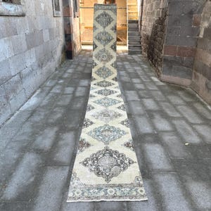 May include: A long, rectangular, vintage-style rug with a cream-colored background and intricate patterns in shades of brown, blue, and beige. The rug is placed on a stone surface, showcasing its length and design.