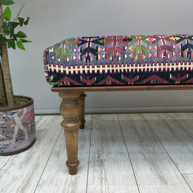 May include: A rectangular bench with a patterned top and wooden legs. The top features a colorful geometric design in shades of blue, red, green, and yellow. The legs are turned and made of light-colored wood.