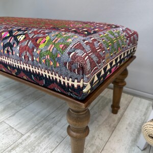 May include: A rectangular ottoman with a patterned textile top in shades of red, gray, and green, resting on four turned wooden legs. The textile features geometric and floral designs.